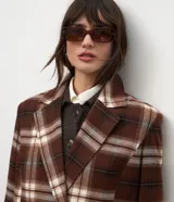Trench coat largo de corte clásico con estampado de cuadros en tonos marrones y beige. Presenta cuello con solapas, cierre frontal con botones, mangas largas y bolsillos laterales aplicados.
