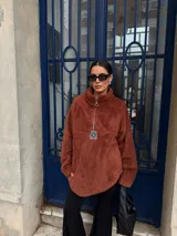 Buzo de corderito color marrón, con cuello alto y cierre frontal parcial. Presenta un diseño holgado con mangas largas y un parche decorativo en el frente.