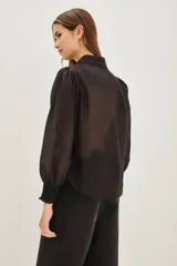 Camisa negra de manga larga con diseño de hombros abullonados y puños elásticos. Presenta un cuello clásico y una tapeta frontal con botones, decorada con detalles de encaje floral a lo largo de la abertura.