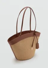 Bolso shopper grande estilo capazo, confeccionado en tejido texturizado efecto rafia en tono beige, combinado con detalles de cuero sintético marrón en los bordes y las asas dobles de hombro. Cuenta con cierre de cremallera superior y un charm colgante decorativo.