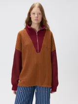 Pullover de tejido acanalado grueso con cuello alto y cierre frontal parcial. Presenta un diseño de bloques de color, con cuerpo en tono beige y mangas en contraste con rayas verticales en tonos rojizos. Incluye un detalle bordado en el pecho.