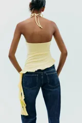 Top negro de cuello halter con lazada, escote recto y hombros descubiertos. Presenta un volante asimétrico en el lateral.