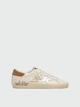Zapatillas Golden Goose modelo Super-Star de cuero napa color blanco, con la icónica estrella lateral laminada en plateado, talón de gamuza marrón y efecto desgastado artesanal. Presenta inscripciones y detalles escritos a mano en los laterales y la lengüeta.