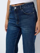 Jean de tiro alto con corte bootcut, confeccionado en denim azul oscuro. Presenta diseño de cinco bolsillos, cierre frontal con botón y cremallera, y terminación con tajos laterales en el ruedo.