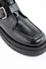Bota corta de cuero negro con suela track dentada y dos correas decorativas con hebillas metálicas en el lateral. Presenta un diseño de puntera cuadrada y cierre lateral.