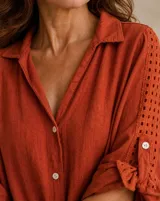 Camisa de lino color terracota, de corte holgado, con cuello camisero, cierre frontal de botones y mangas largas con detalles perforados y trabilla para arremangar.
