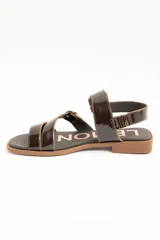 Sandalias de cuero charolado negro con dos tiras con hebillas metálicas plateadas y tira en el talón. Suela plana color marrón.
