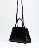 Cartera de mano de símil cuero color negro, con diseño estructurado, doble asa rígida y correa ajustable desmontable. Cuenta con costuras verticales decorativas en el frente, tiras laterales con broches y el logo metálico de la marca en la esquina inferior.