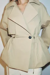 Trench corta de color beige con cuello de solapa y manga larga. Cuenta con bolsillos delanteros, cintura ajustable con cordones, bajo abullonado y cierre frontal cruzado con un botón visible. El interior está forrado con tela a cuadros.