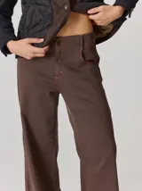 Pantalón de corte relajado color marrón, confeccionado en una mezcla de algodón y elastano. Presenta costuras en contraste color rojo en bolsillos y bragueta, con cierre de botón metálico.