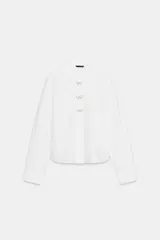Camisa de popelín blanca con cuello solapa y manga larga. Presenta detalle de botones joya decorativos en forma de lazo en la parte frontal y cierre oculto por solapa.