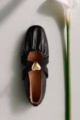 Balerina de cuero ovino color negro, con diseño fruncido en la puntera y tiras elásticas cruzadas sobre el empeine adornadas con una aplicación metálica en forma de corazón dorado.
