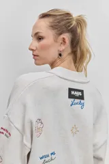 Cardigan de punto azul con cuello en V, cierre frontal de botones y bolsillos delanteros. Presenta bordados decorativos con motivos variados y frases en colores contrastantes.