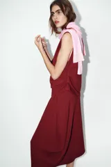 Vestido largo color borgoña, con escote barco y manga caída/sisa. Presenta un drapeado lateral que genera un bajo asimétrico.