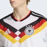 Camiseta de fútbol Adidas de la primera equipación de Alemania, modelo 26. Diseño principal blanco con un patrón en V en el pecho que incorpora los colores de la bandera alemana (negro, rojo y amarillo/dorado). Posee cuello en V con ribete rojo y detalles en las mangas. El escudo de la federación alemana con cuatro estrellas está bordado en el pecho, junto al logo de Adidas.