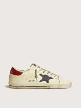 Zapatillas Golden Goose modelo Super Star, color crema con detalles en azul y rojo. Diseño vintage con efecto desgastado, estrella azul en el lateral y contrafuerte rojo.