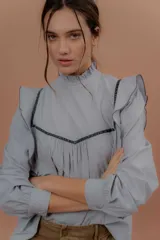 Blusa de jeans fino color celeste con detalles de terciopelo negro en contraste. Presenta cuello alto con volados, mangas largas abullonadas con puños ajustados y volados decorativos en los hombros y el frente.