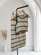 Conjunto de dos piezas de tejido crochet, color beige claro con ribetes negros. Incluye un top tipo musculosa de corte cropped y una falda larga ajustada. El tejido presenta un patrón de ondas o escamas.