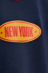 Camiseta de cuello redondo y manga corta, color azul marino con ribetes en color rojo en cuello y mangas. Presenta un estampado frontal ovalado con el texto New York en color naranja.