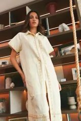 Vestido camisero de lino color lila con bordados rojos en contraste. Presenta un diseño de largo midi con cuello camisero, cierre frontal de botones y mangas cortas. Los detalles bordados recorren la botonadura, los puños y el ruedo de la prenda.