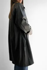 Trench de ecocuero negro de corte relajado y largo medio. Presenta cierre frontal con botones, bolsillos delanteros funcionales y tajo posterior. Las mangas cuentan con un forro interno a rayas que queda visible al doblarlas.