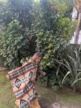 Poncho estilo navajo confeccionado en mezcla de lana y algodón, con estampado geométrico multicolor en tonos beige, marrón, negro, turquesa y blanco, y terminación de flecos en el ruedo.