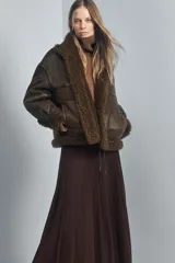 Chaqueta tipo bomber de cuello alto y manga larga, confeccionada en material con efecto cuero envejecido y forro interior de pelo sintético. Presenta cierre frontal con botones, bolsillos delanteros y bajo ajustable con cordones.