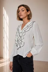 Blusa blanca de manga larga con escote en V y volados frontales con reborde negro. Presenta mangas abullonadas con puños elásticos y corte fluido.