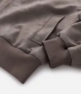 Campera de material sintético en color gris topo, con cuello inglés, cierre frontal por cremallera, mangas largas con puños elásticos y diseño de relieve tribal en la espalda.