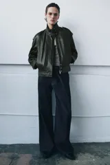 Chaqueta bomber de piel color verde oscuro, con cuello alto, manga larga con puño, bolsillos delanteros y trabillas en los hombros. Cierre frontal con cremallera y botones ocultos por solapa.