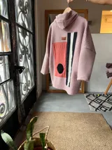 Campera con capucha confeccionada en tejido tipo corderito color rosa, con diseño de collage geométrico en el frente que incluye bloques de color en naranja, negro y blanco. Presenta mangas largas de corte holgado y diseño artesanal único.