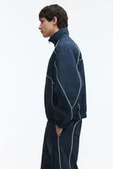 Campera deportiva de nylon en color verde oscuro con estampado a cuadros, cuello elevado, cierre frontal y ribetes blancos en contraste. Cuenta con elástico en puños y bajo, y forro de malla.