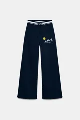 Pantalón de felpa azul marino con tiro alto, cintura elástica con cordón ajustable y diseño de pierna ancha. Presenta un estampado frontal con el logo de St. Moritz y un sol amarillo.