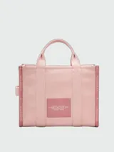 Bolso tipo tote bag de Marc Jacobs en color beige, confeccionado en lona resistente. Presenta el texto "THE TOTE BAG" y "MARC JACOBS" impreso en la parte frontal en color negro. Cuenta con asas superiores y una correa de hombro ajustable y extraíble, además de ribetes laterales negros con inscripciones.