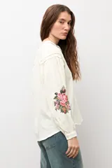 Camisa de color natural con mangas largas abullonadas y bordado floral multicolor en los puños. Presenta cuello mao y cierre frontal con botones.