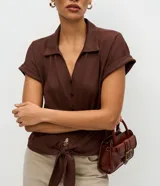 Blusa color marrón con cuello solapa, mangas cortas con vuelta y cierre frontal de botones. Cuenta con un diseño texturizado y un lazo para anudar en el bajo.