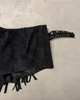 Short pollera negro de efecto gamuza o cuero sintético, con borde festoneado y tachas metálicas plateadas. Presenta flecos largos en el ruedo inferior y se ajusta con una hebilla en el lateral.