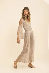 Vestido largo color champagne, con tejido de punto calado y textura brillante. Presenta escote en V, cintura marcada con frunces y mangas largas con abertura tipo capa.