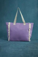 Bolso tipo tote lila con flecos blancos en los laterales y asas de tela color crudo.