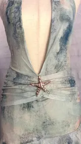 Vestido corto de microtul celeste con efecto tie-dye en tonos verde agua y gris. Diseño halter con escote profundo en V, fruncido en la cintura con un aplique de estrella de mar brillante, y ruedo con volado.