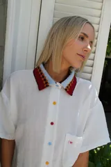 Camisa blanca de lino de manga corta, con cuello tejido a crochet en tonos rojo, marrón y celeste con patrón geométrico. Presenta botones de colores surtidos en el frente y un pequeño parche bordado con un corazón rosa en el bolsillo del pecho.