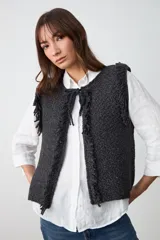 Chaleco de tejido de punto grueso en color negro, con diseño sin mangas, textura irregular y detalle de flecos en los bordes frontales. Cuenta con un cierre de lazo en la parte superior del cuello.