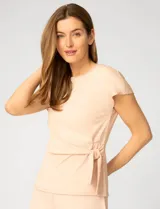 Remera de manga corta color beige, confeccionada en mezcla de modal, con cuello redondo y detalle de lazo lateral que genera un drapeado natural en la cintura.