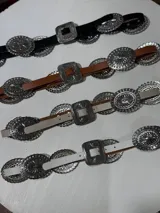 Cinturón marrón de cuero con hebilla plateada rectangular con diseño de flores y apliques metálicos circulares con diseño de flores.