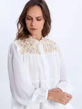 Camisa blanca vaporosa con cuello mao, botonadura frontal oculta y mangas largas abullonadas con puños elásticos. Presenta un diseño de paneles fruncidos y detalles bordados con motivos geométricos dorados en la parte superior delantera.