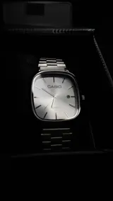 Reloj Casio con caja cuadrada de bordes redondeados y malla de acero inoxidable. Presenta una esfera plateada con marcadores de hora lineales, manecillas finas y ventana de fecha en la posición de las tres.