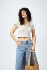 Remera corta de punto acanalado color beige claro, con escote en V fruncido y bordeado con puntilla verde claro. Tiene mangas cortas y un calce ajustado al cuerpo.