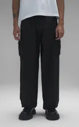 Pantalón cargo de estilo técnico en color negro, confeccionado en textil memory con acabado sutilmente brillante. Presenta una silueta oversize, cinturón regulable integrado con hebilla de encastre, dos bolsillos laterales, dos bolsillos cargo de gran tamaño y un bolsillo trasero.