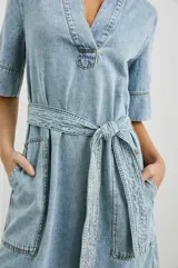 Vestido camisero corto de denim celeste con cuello en V, mangas cortas y cinturón de lazo. Presenta bolsillos laterales y pespuntes a contratono.