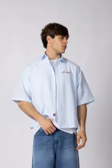 Camisa de manga corta de popelina rayada, con calce boxy y bordado sutil en el pecho. Presenta cierre frontal con botones y diseño abierto.